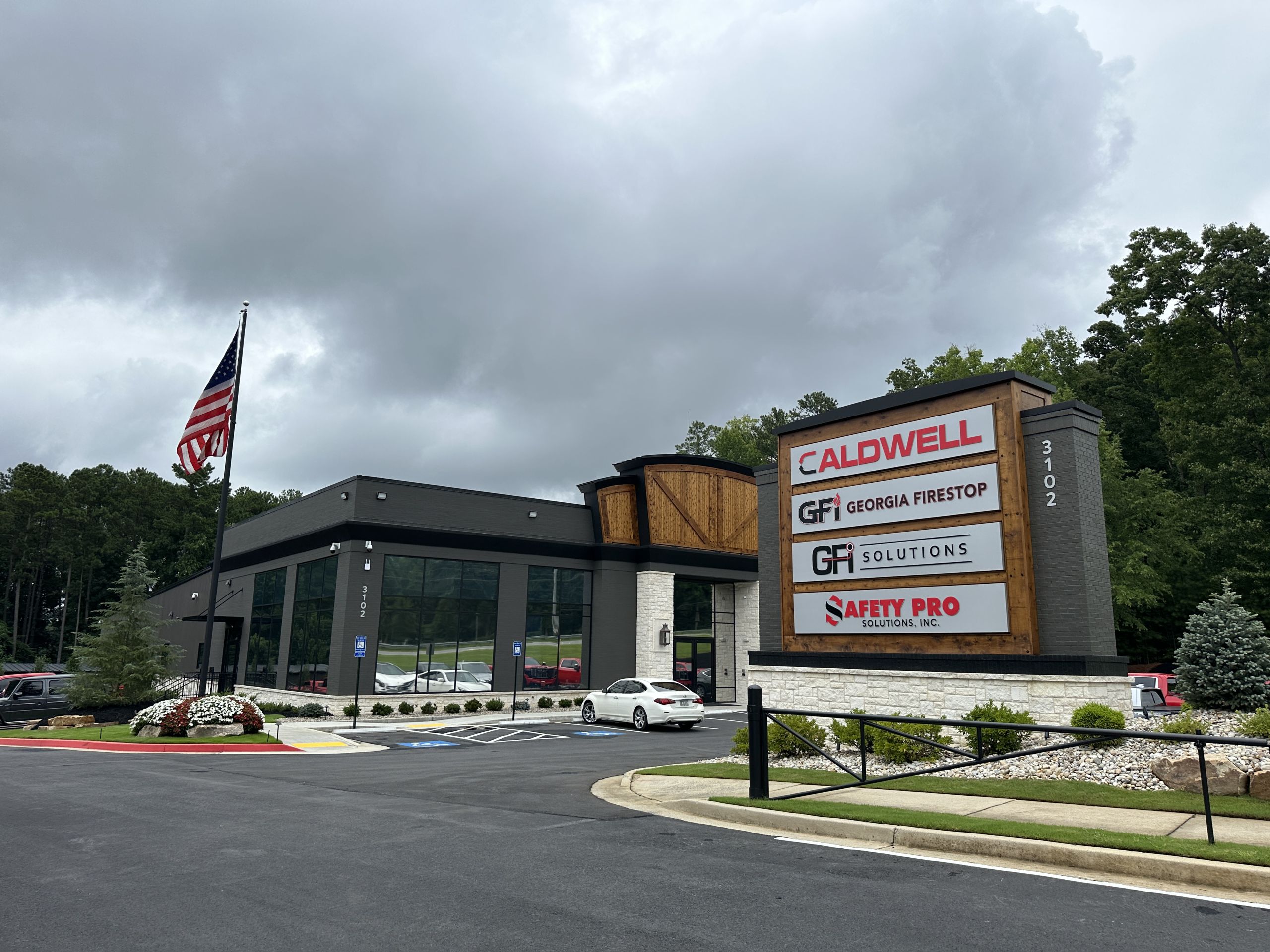 Modern commercial building with large windows, an American flag, and a sign listing Caldwell, Georgia Firestop, GFI Solutions, and Safety Pro. This impressive new headquarters highlights the growth of local businesses in the area. Trees and cloudy sky form a peaceful background.